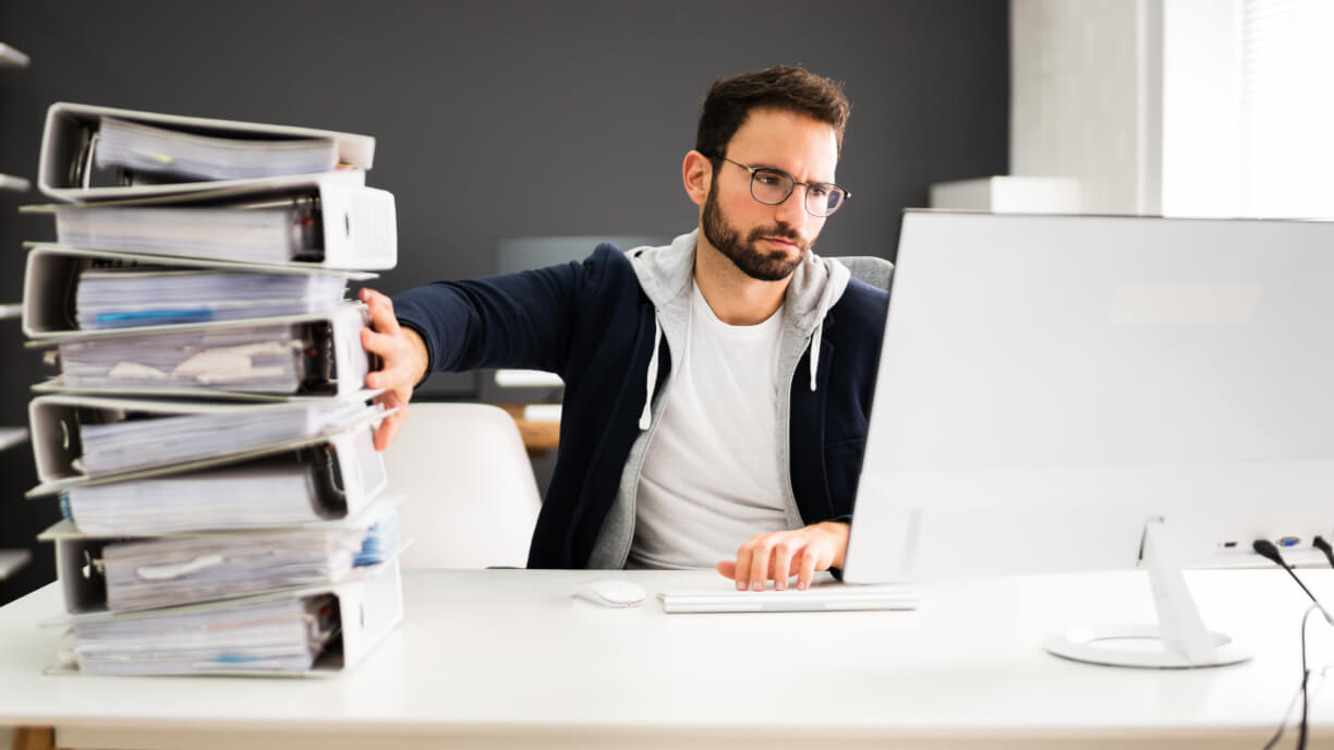 A man sitting at a desk using management software to organize a stack of folders in front of him.